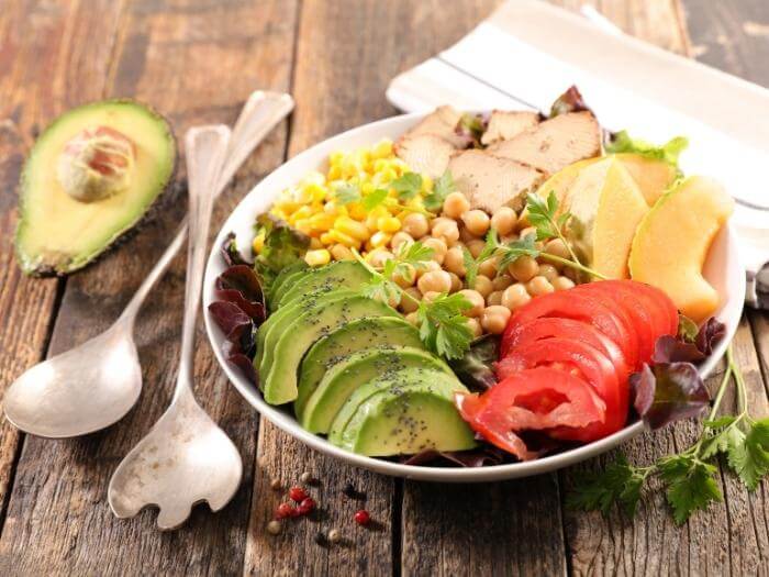 Bowl meal with half sliced avocado, sliced tomatoes, papaya, tofu, corn,chick-pea on a white bowl on a wooden table