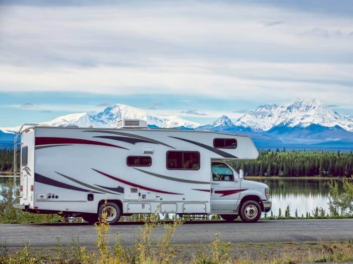A class C motorhome parked by the side of the road with a beautiful lake and aspen trees and ice capped mountain view.