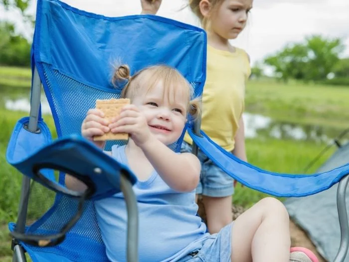 a toddler eating a cracker in a camping chair with another toddler behind