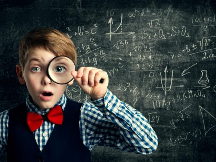 A boy with magnifying glass with black board formulas at the back