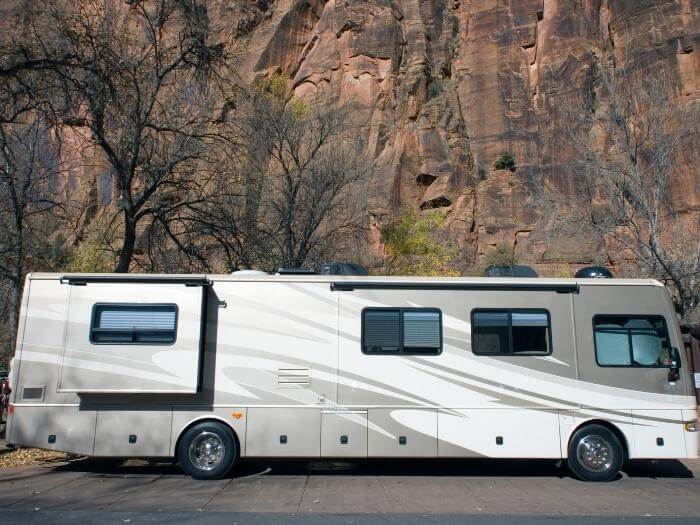 A gray and white Class A RV by a mountain side road with a few trees behind.