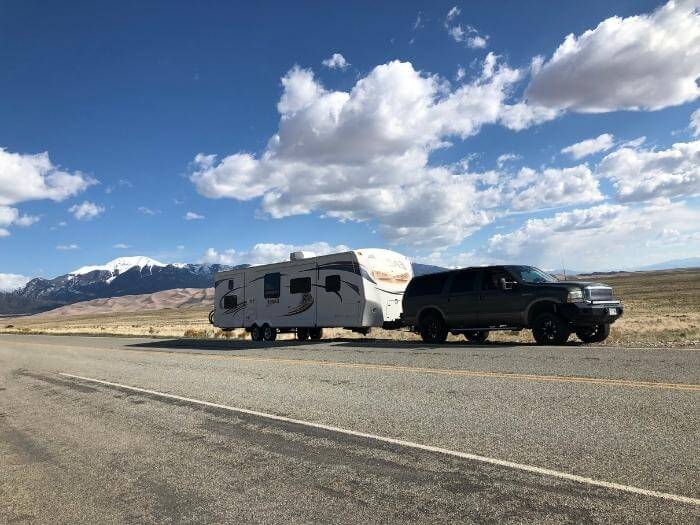 A 35 feet travel trailer pulled by an SUV by the side of the road on a clear day cloudy sky.