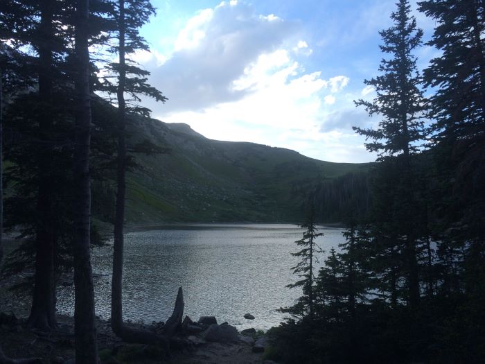Photo of a lake at dusk, with tall pine trees in the foreground and mountains rising behind it