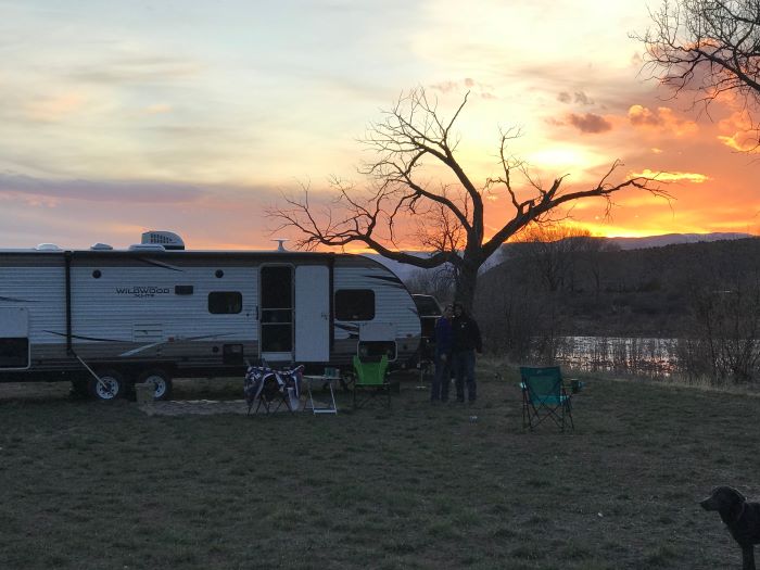 A couple and their dog stand outside their camper next to a river at dusk.