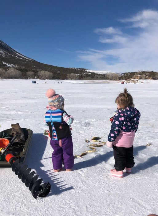 Two young girls in snow gear stand near an ice auger and pile of perch while ice fishing on a frozen lake