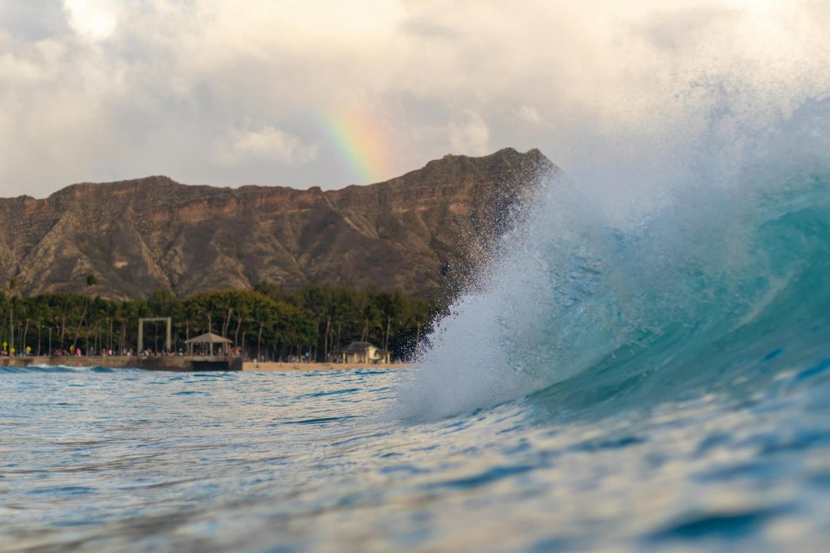Ocean wave curling with a rainbow visible over Diamond Head in Waikiki, Hawaii